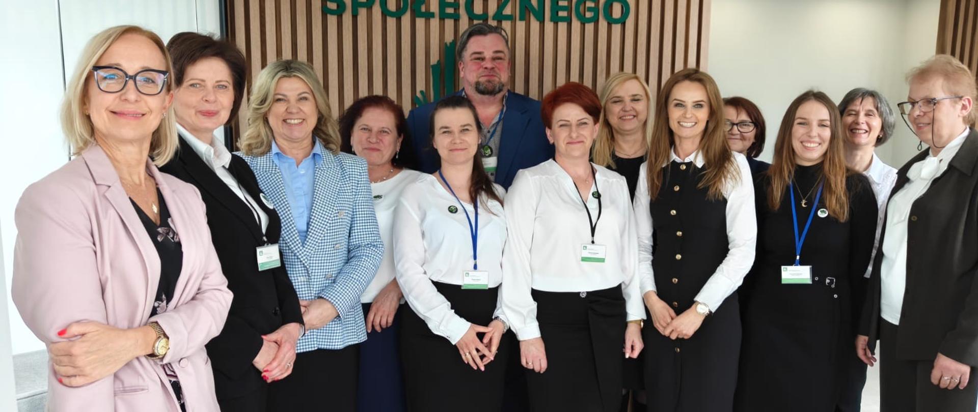 Grupa osób ubranych elegancko pozuje do zdjęcia, w tle duże zielone logo KRUS, które znajduje się na ściance z drewnianych lameli.
