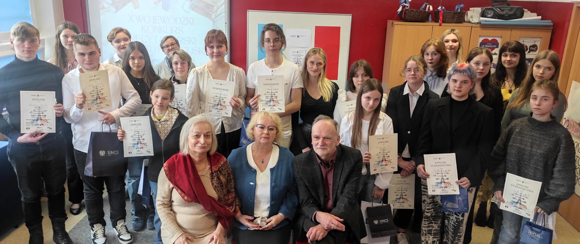 Sala szkolna. Na pierwszym planie siedzą trzy starsze osoby; za nimi grupa kilkunastu młodych osób stoi, większość trzyma w rękach dyplomy
