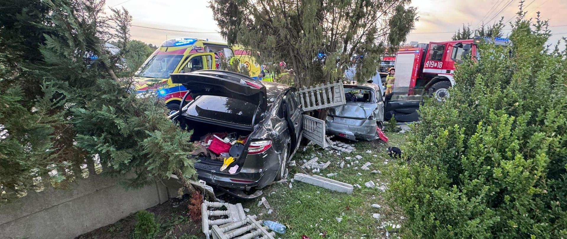 Rozbite dwa samochody osobowe oraz betonowe ogrodzenie przy prywatnej posesji. W tle karetka pogotowia oraz samochód ratowniczo-gaśniczy. 