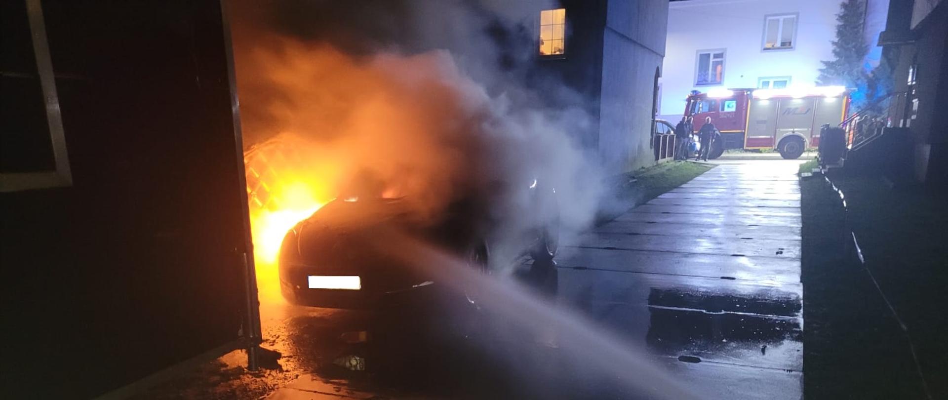 Na zdjęciu widoczny jest samochód osobowy zaparkowany przy budynku. Prawy przód samochodu jest objęty pożarem. Na zdjęciu widoczny podany prąd wody w celu ugaszenia pożaru. W oddali widać stojący samochód pożarniczy koloru czerwonego. Pora dnia na zdjęciu - wieczór.