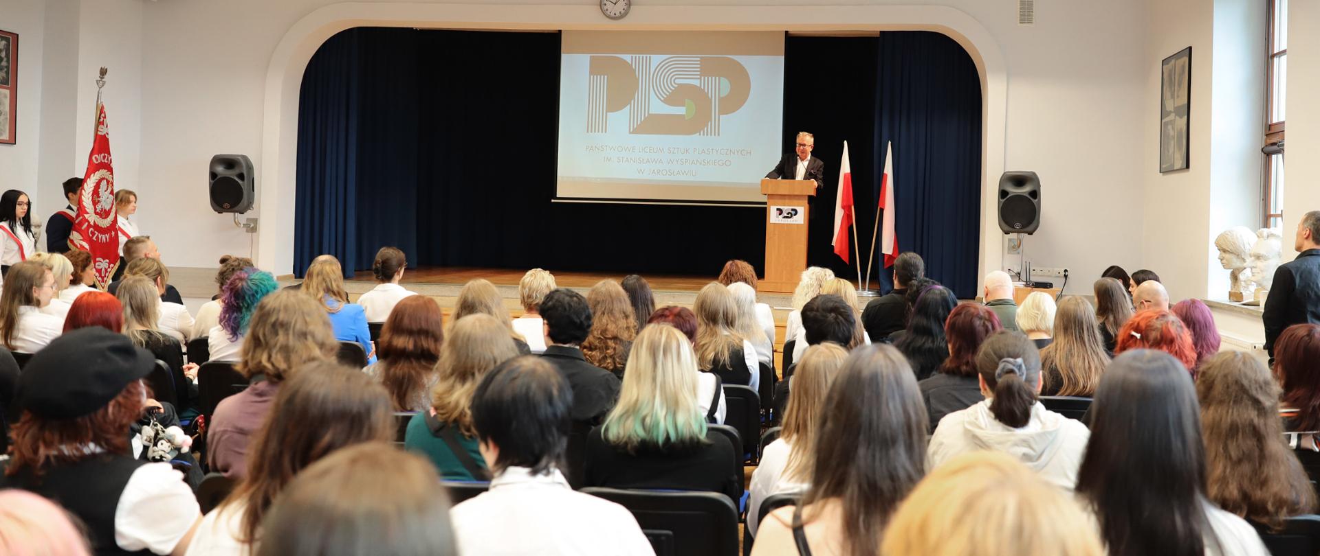 Fotografia przedstawia scenę, na której stoi i przemawia mężczyzna. Po lewej sztandar i 3 osoby. Na pierwszym planie wiele osób oglądających wydarzenie.