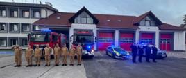 Strażacy i policjanci stoją przed samochodami służbowymi i oddają cześć strażakom, którzy zginęli w pożarze w Nisku. Strażacy i policjanci są w umundurowaniu koszarowym, stoją na baczność przed bramami garażowymi Komendy Powiatowej PSP w Lubaniu. Pojazdy mają włączone sygnały świetlne.