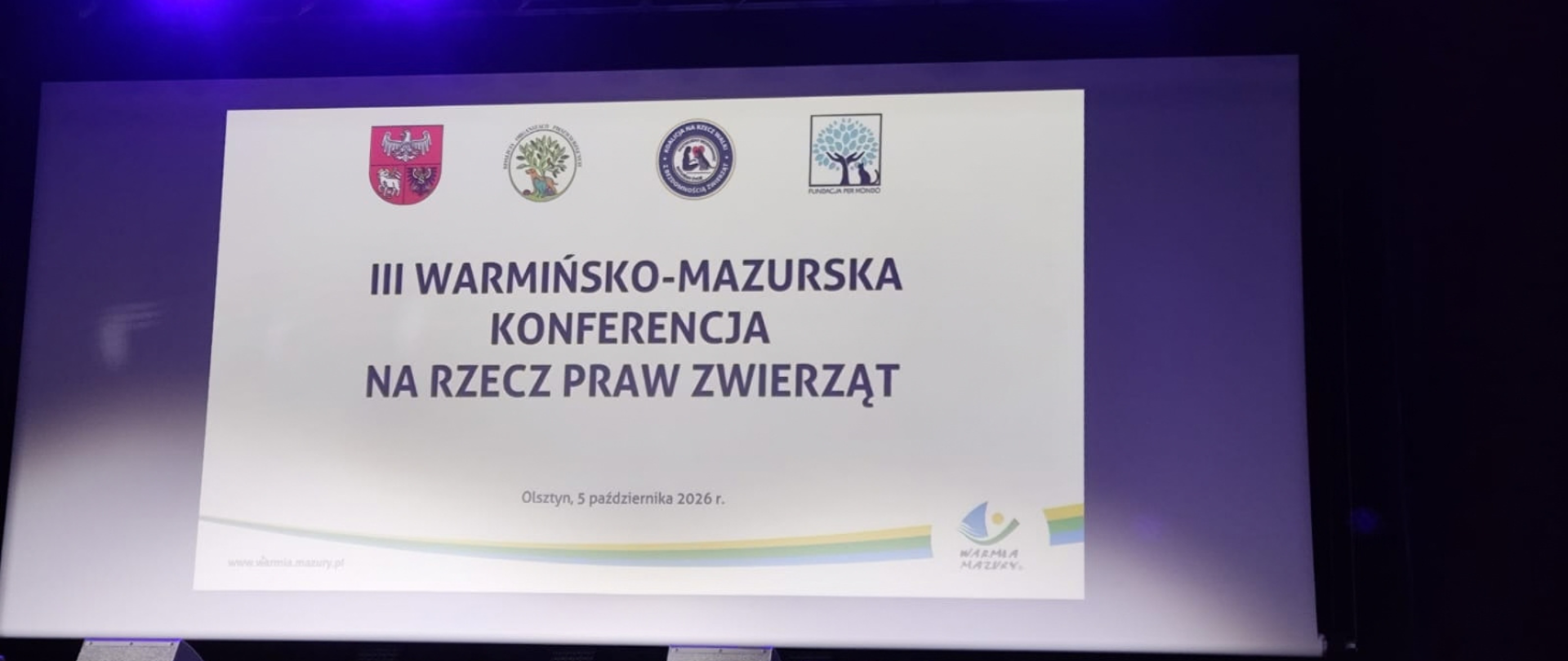 Zdjęcie ekranu podczas wydarzenia. Na slajdzie widnieje tytuł „III Warmińsko-Mazurska Konferencja na Rzecz Praw Zwierząt”, data „Olsztyn, 5 października 2026 r.” oraz logotypy organizatorów i partnerów