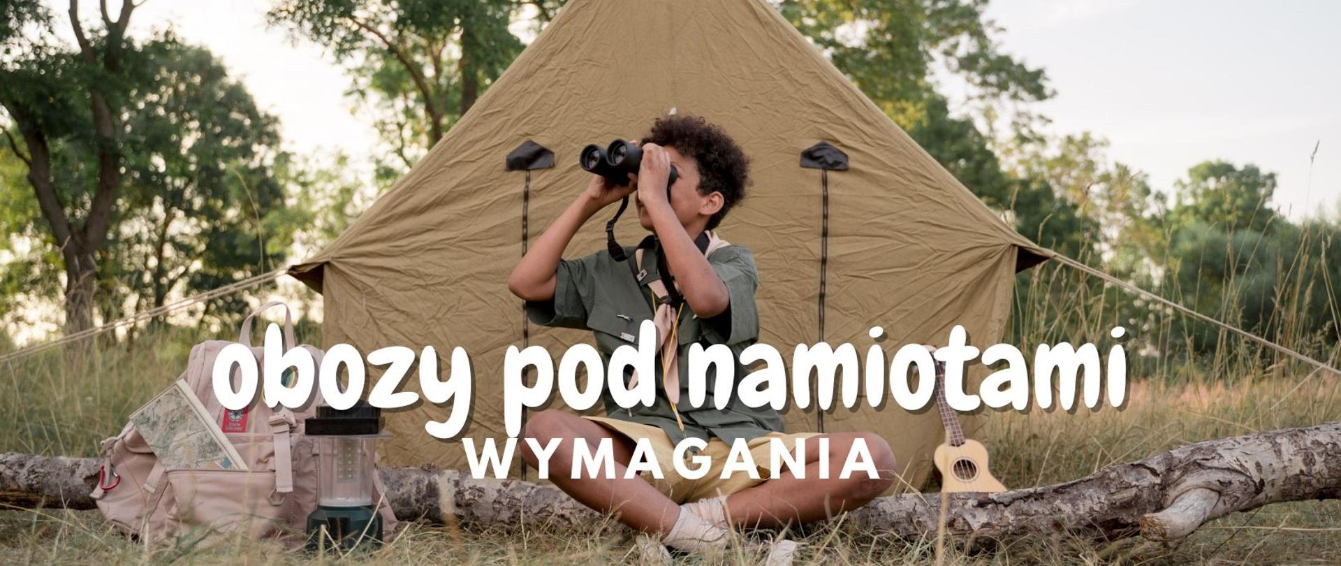 Chłopiec siedzący przed skautowskim namiotem trzymający lornetkę