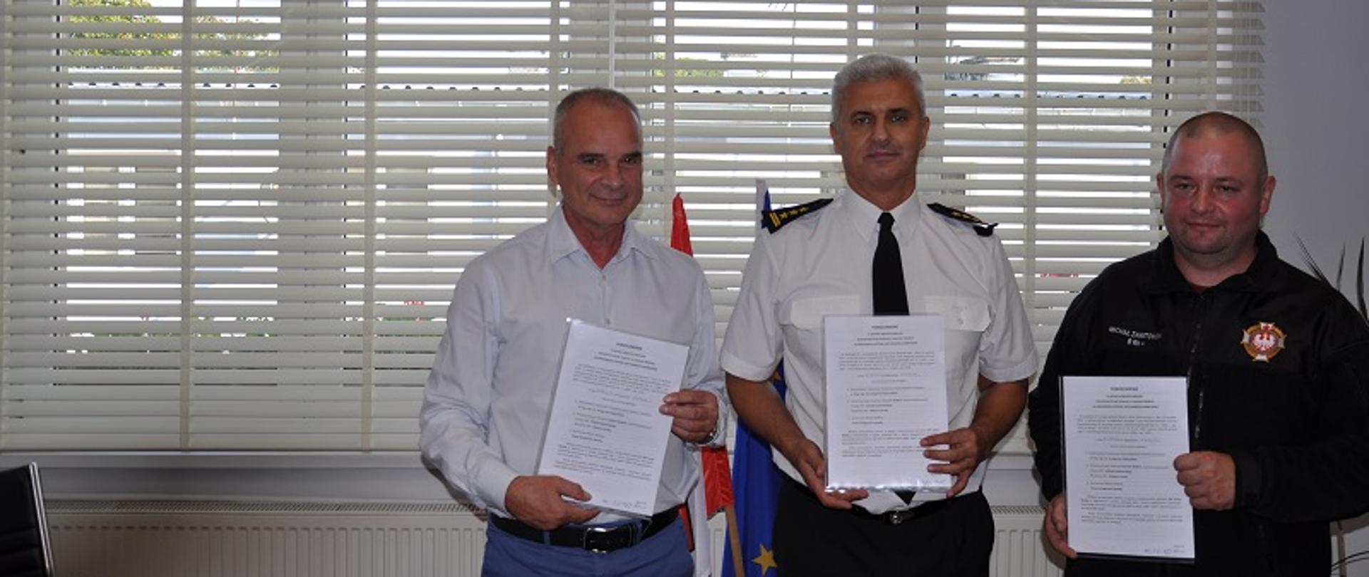 Zdjęcie przedstawia akt podpisania porozumienia o włączeniu jednostki OSP Lesiny Wielkie do krajowego systemu ratowniczo gaśniczego. Zdjęcie obrazuje gabinet burmistrza miasta Wielbark. Na zdjęciu znajdują się następujące osoby burmistrz Grzegorz Zapadka, komendant powiatowy PSP w Szczytnie Grzegorz Rybaczyk oraz Prezes OSP Lesiny Wielkie druh Michał Zawitowski.