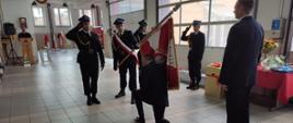 W garażu w strażnicy poczet sztandarowy strażaków pochyla sztandar, strażak klęka na prawe kolano całuje płat sztandaru, drugi strażak stoi przed sztandarem.