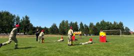 Pokaz grupy Młodzieżowej Drużyny Pożarniczej z Białej Rawskiej. Czwórka dzieci podzielona na dwójki jeden leży na trawie i w dłoni trzyma odcinek wężowy z wodą, celujących w tabliczkę, a drugi za nim pompuje zbiornik z wodą.