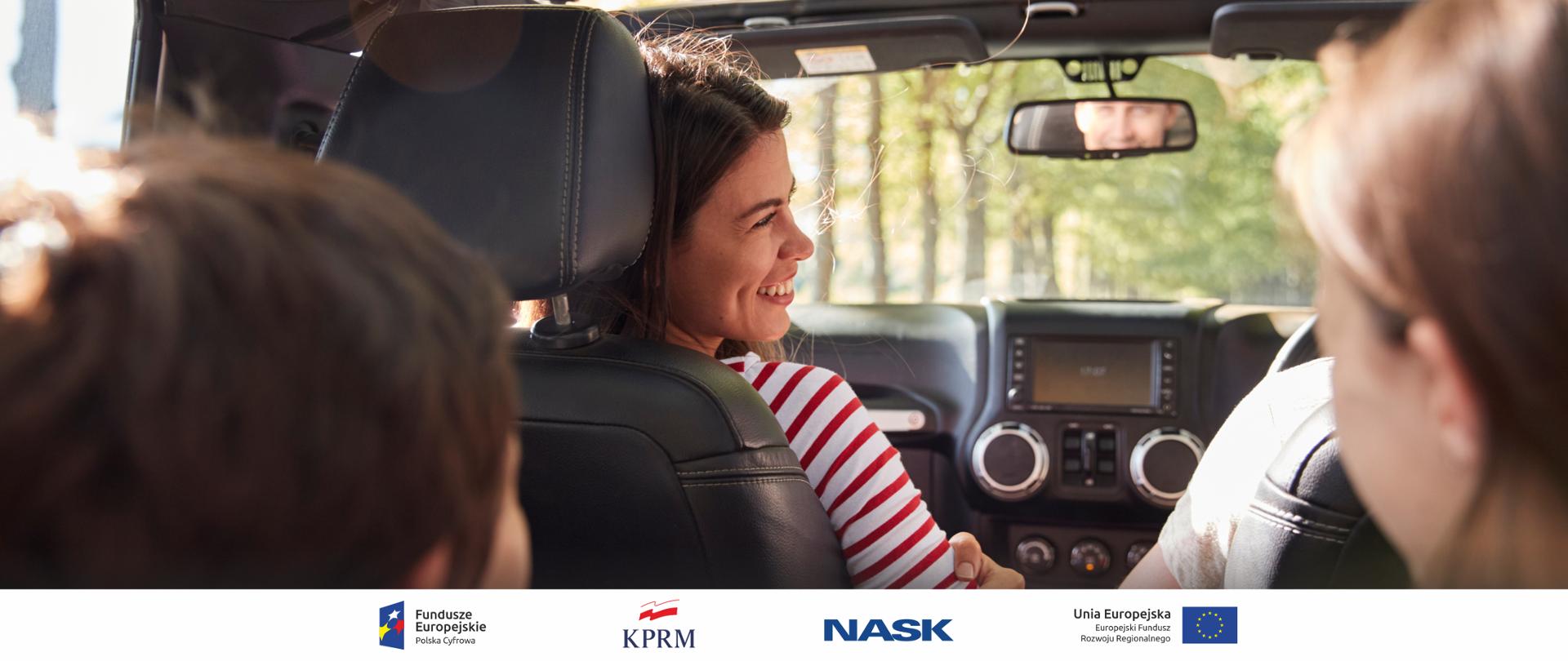 Zdjęcie wnętrza samochodu. Za kierownicą siedzi uśmiechnięta młoda kobieta. W przednim lusterku widać mężczyznę. Na tylnym siedzeniu widać głowy dzieci. 