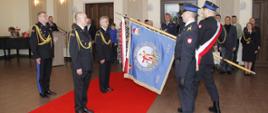 Sala w Urzędzie miasta Brzeziny na zdjęciu poczet sztandarowy komendy powiatowej psp w brzezinach przed pochyloną flagą stoją ustępujący komendant psp w brzezinach starszy brygadier dariusz guzek oraz komendant powołany na pełniącego obowiązki komendanta powiatowego psp w brzezinach za nimi toarzyszący komendant wojewódzki nadbrygadier Grzegorz Janowski