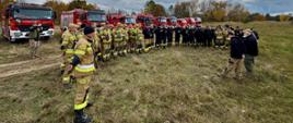Zbiórka strażaków w terenie piaszczystym gdzie mają ćwiczyć przemieszczanie się pojazdami pożarniczymi w trudnym terenie. W tle ustawione wozy strażackie.