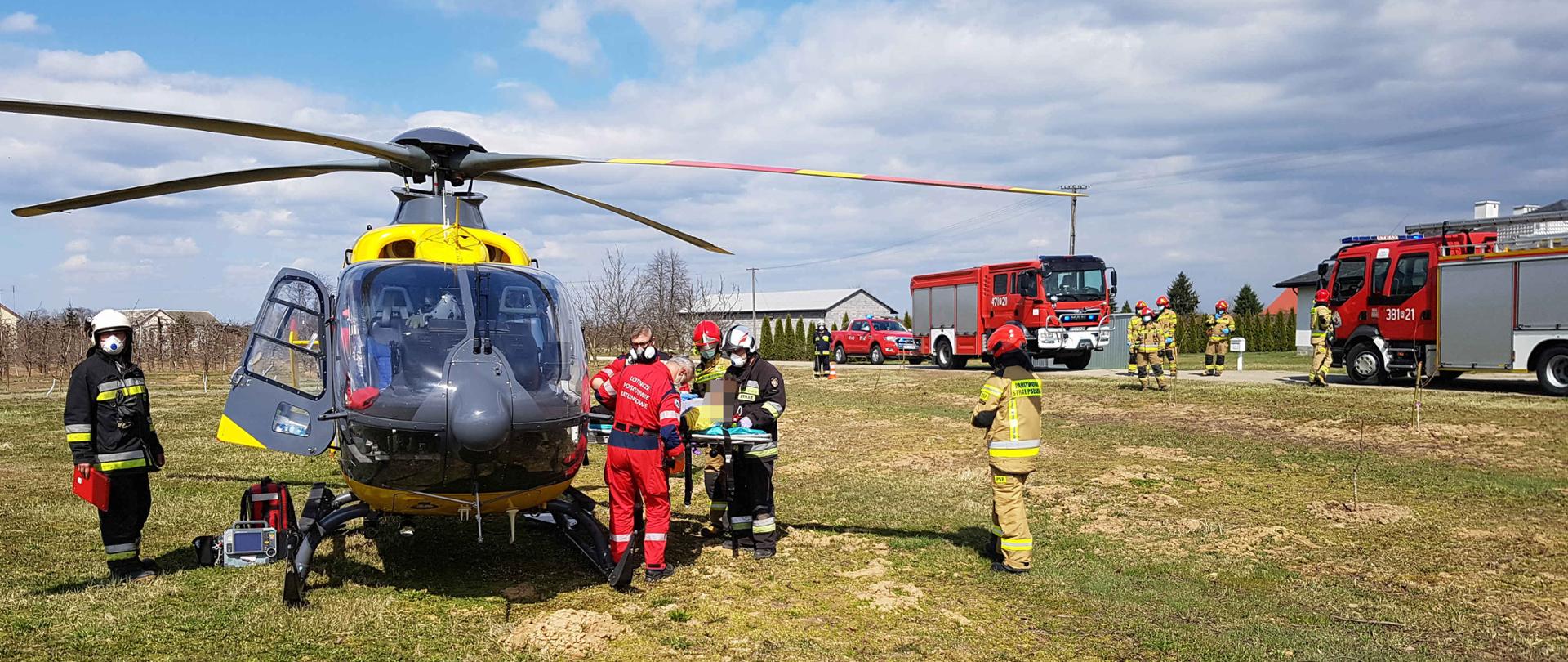 Zdjęcie zrobione w dzień. Na zdjęciu widać śmigłowiec LPR, który stoi na miejscu lądowania. Obok śmigłowca LPR widać ratowników medycznych i strażaków, którzy pomagają wejść do śmigłowca LPR osobie poszkodowanej. W oddali widać trzy samochody strażackie oraz strażaków, którzy zabezpieczają lądowisko śmigłowca. 