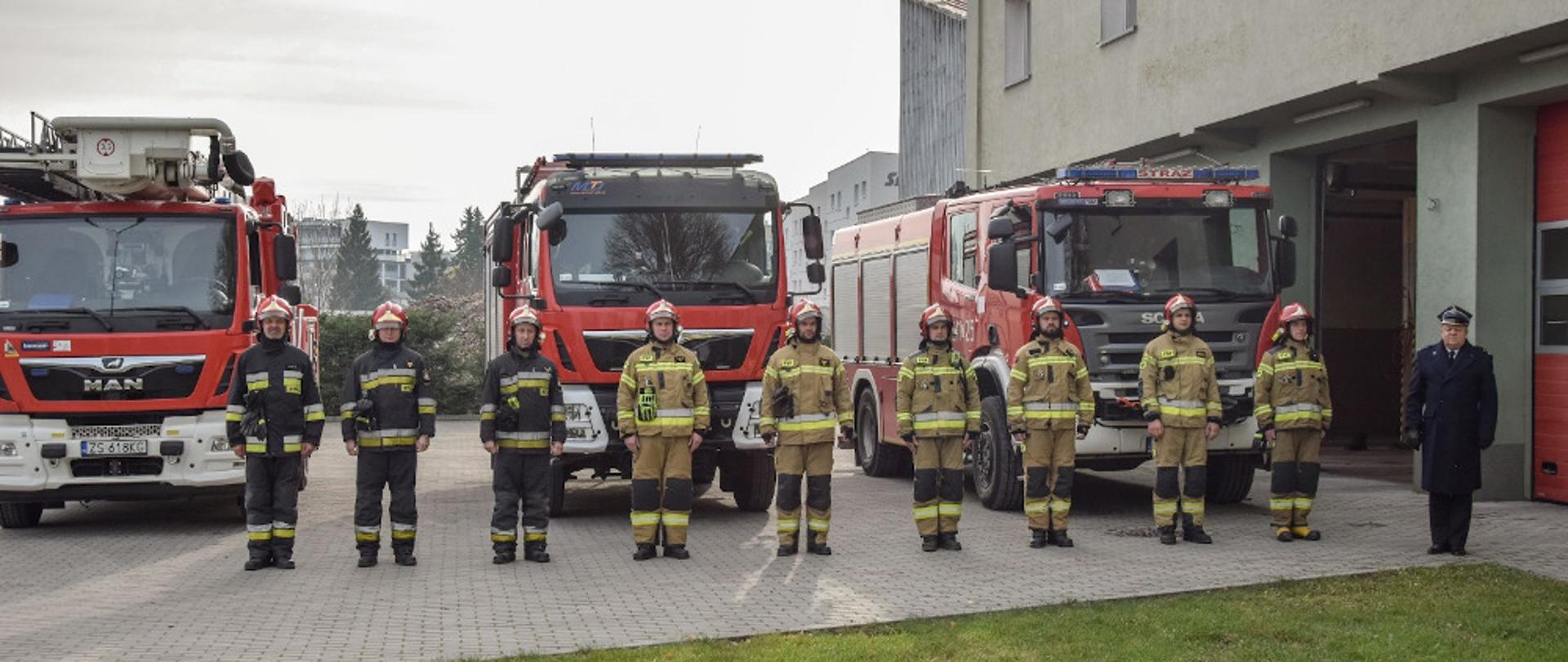 Strażacy ubrani w ubrania specjalne oraz komendant powiatowy bryg. Andrzej Wójtowicz w płaszczu wyjściowym zimowym stoją na tle samochodów ratowniczo-gaśniczych 