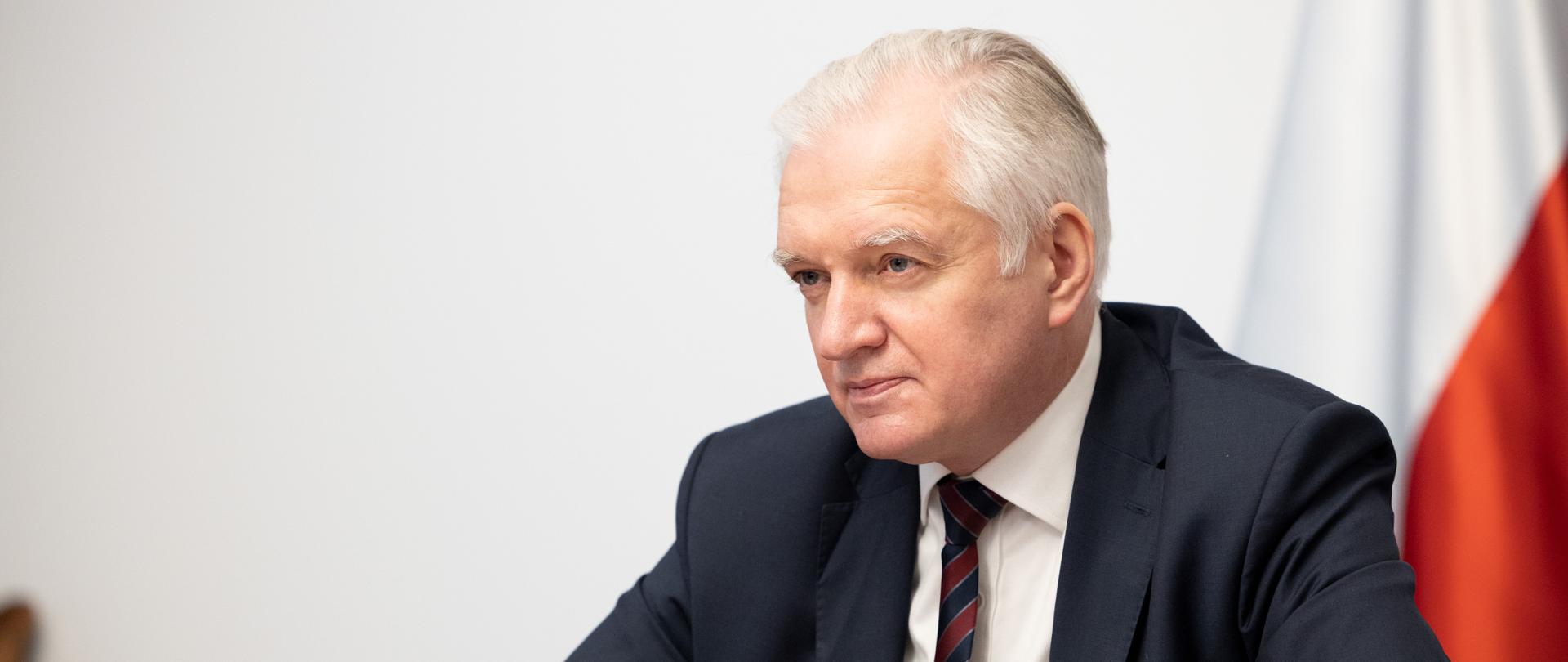 Jarosław Gowin przy stole konferencyjnym, za nim flagi, przed nim mikrofon. Widok z boku.
