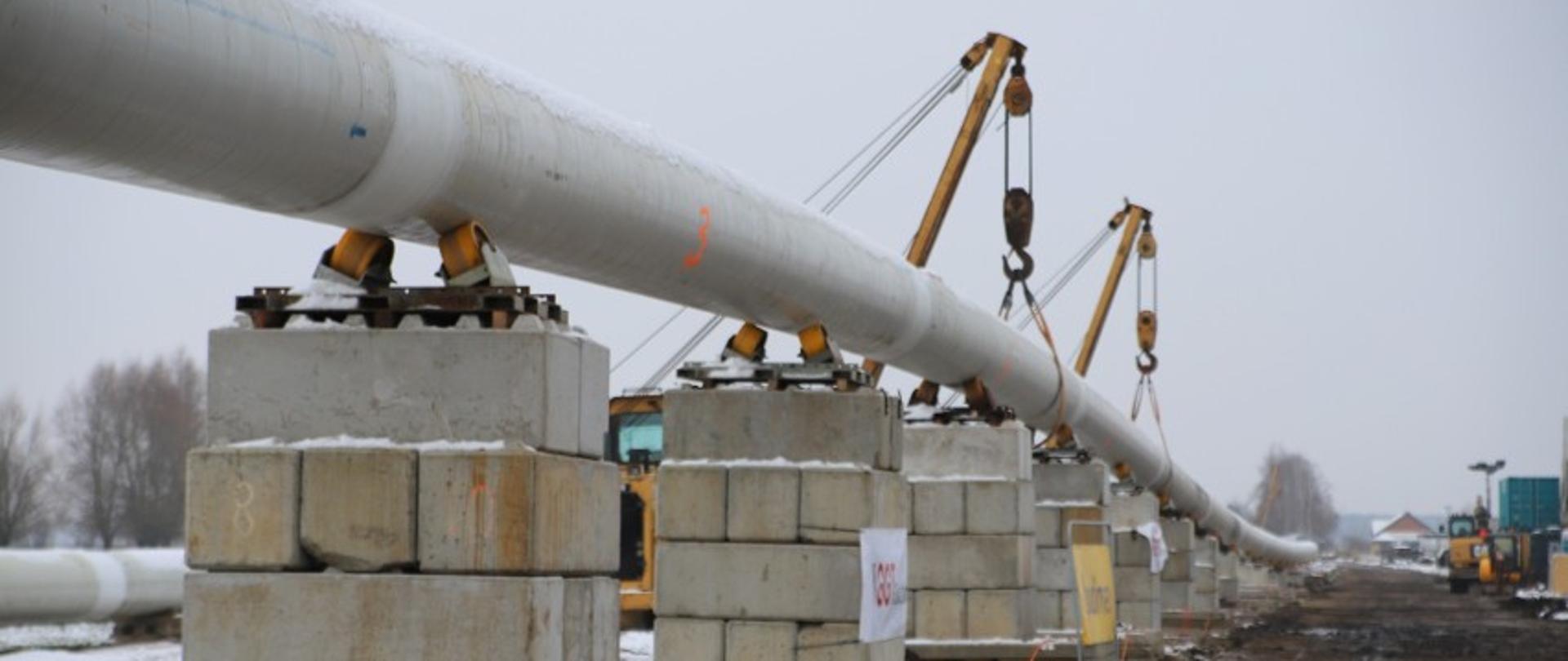 Dług szara rura oparta o betonowe bloczki, w oddali haki koparek, podtrzymujących rurę.