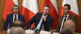 Przy drewnianym stole siedzi minister Czarnek i mówi do mikrofonu, obok niego dwóch mężczyzn w garniturach, za nimi polskie i włoskie flagi.