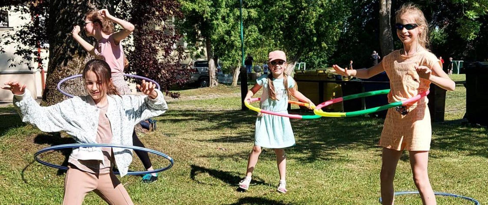 Zdjęcie kolorowe. Cztery dziewczynki w letnich strojach uprawiają gimnastykę na trawniku. W tle zielone drzewa.