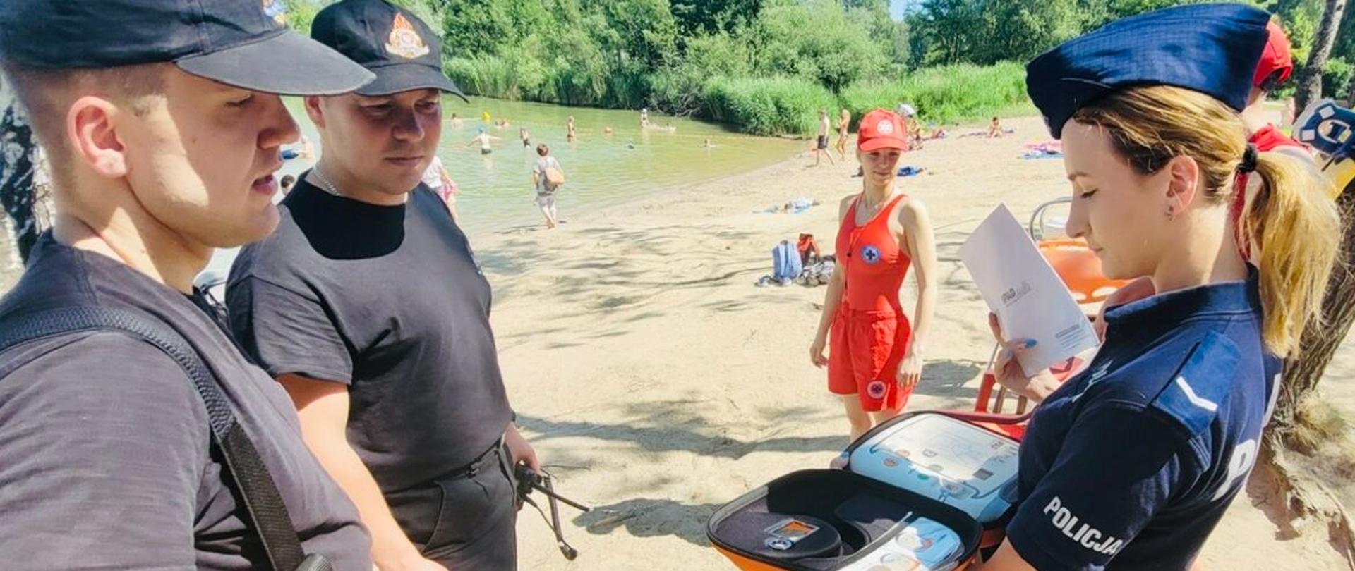 Na zdjęciu na plaży znajduje się Policjantka ubrana w mundur oraz dwóch umundurowanych strażaków.. Na dalszym planie widać ratowniczkę oraz ludzi na plaży. W tle widac drzewa