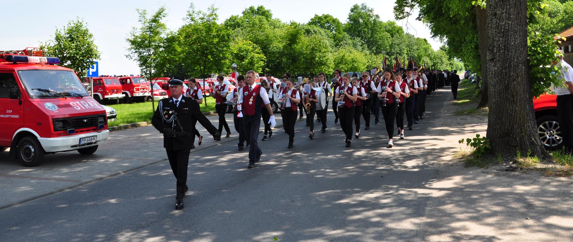 Na zdjęciu widać pojazdy pożarnicze oraz orkiestrę strażacką podczas przemarszu.