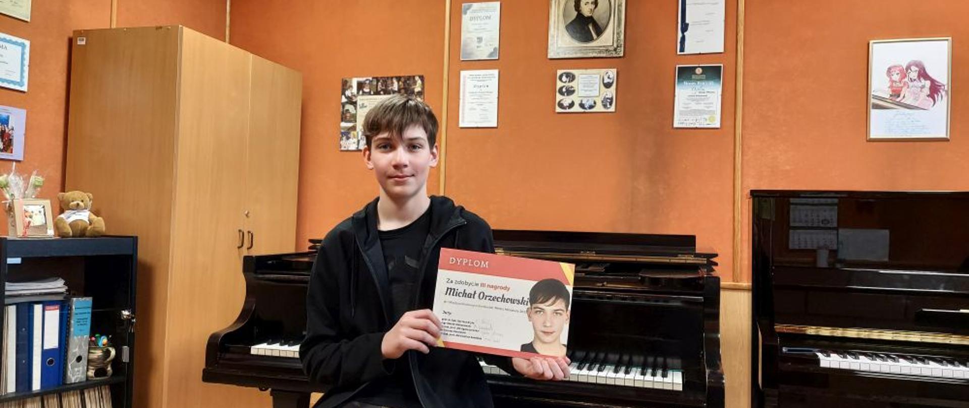 Zdjęcie kolorowe. Laureat konkursu Michał Orzechowski siedzi na tle fortepianu trzymając w ręku dyplom. 
