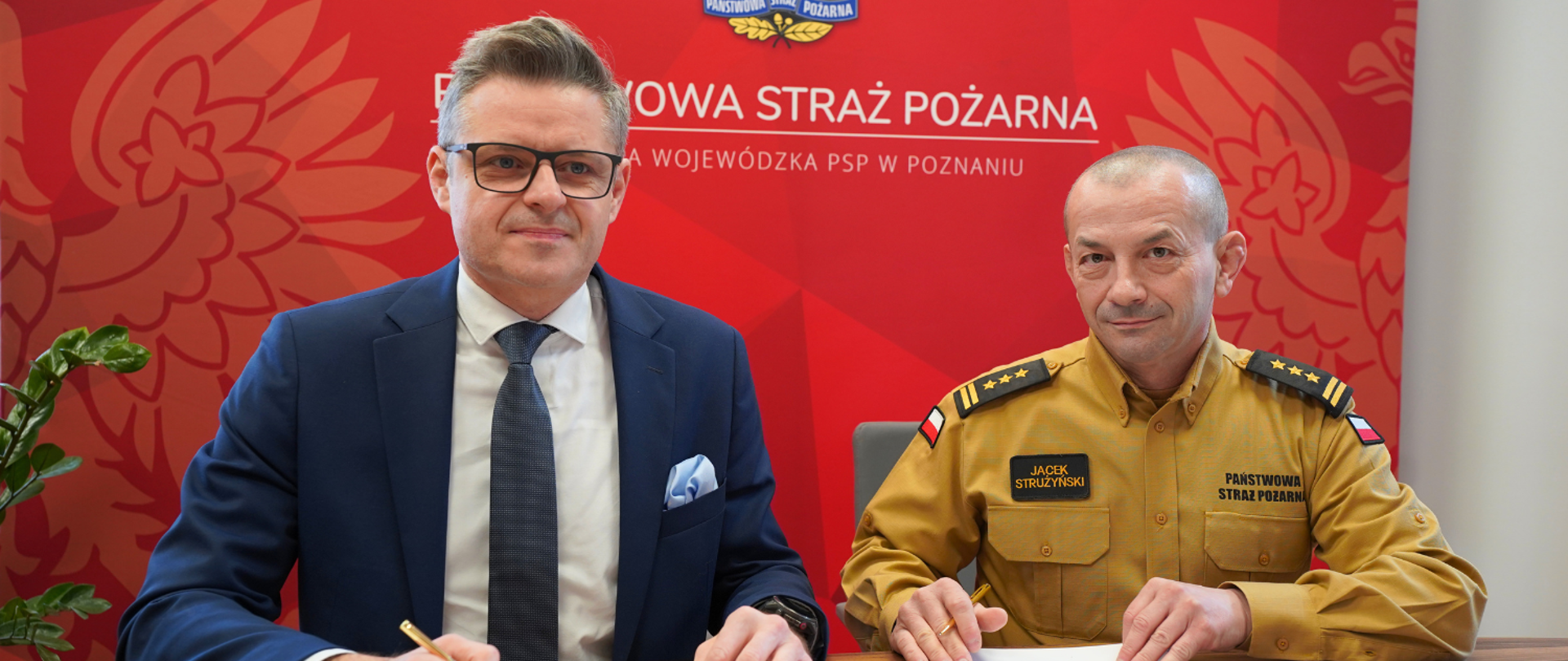 Szef funduszu ochrony środowiska oraz komendant wojewódzki PSP podpisali umowę na dofinansowanie zadań PSP. Panowie podpisują dokumentację na tle ścianki reklamowej PSP w biurze komendy.