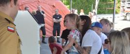 Dzień otwarty Plac wewnętrzny komendy. Pod namiotem, na stole znajduje się mobilny symulator zagrożeń pożarowych. Stojący obok strażak opowiada gościom (rodzicom z dziećmi) o zagrożeniach. W tle namiot pneumatyczny.
