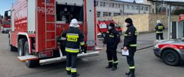 Zdjęcie przedstawia strażaka OSP egzaminowanego przez komisję z obsługi autopompy. Od lewej strony na zdjęciu uwidoczniono: ustawiony tyłem ciężki samochód gaśniczy z otwartym przedziałem autopompy. Przed tablicą rozdzielczą autopompy tyłem do patrzącego stoi strażak OSP w Granatowym ubraniu specjalnym i białym hełmie. W środku kadru na prawo od samochodu stoją dwaj członkowie komisji: strażacy PSP w ubraniach koszarowych, czapkach zimowych i maseczkach. W tle budynek komendy i kolejny strażak OSP obsługujący linię szybkiego natarcia. 