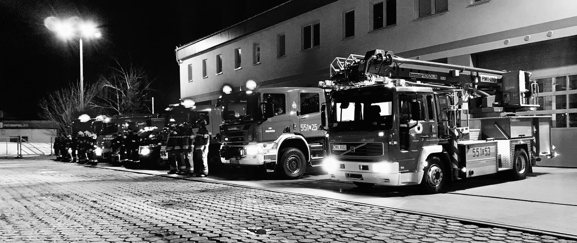 Zdjęcie czarno-białe, na którym na tle budynku komendy powiatowej PSP w Wąbrzeźnie stoją wozy strażackie, oraz strażacy składający hołd ofiarą dzisiejszego wypadku. 