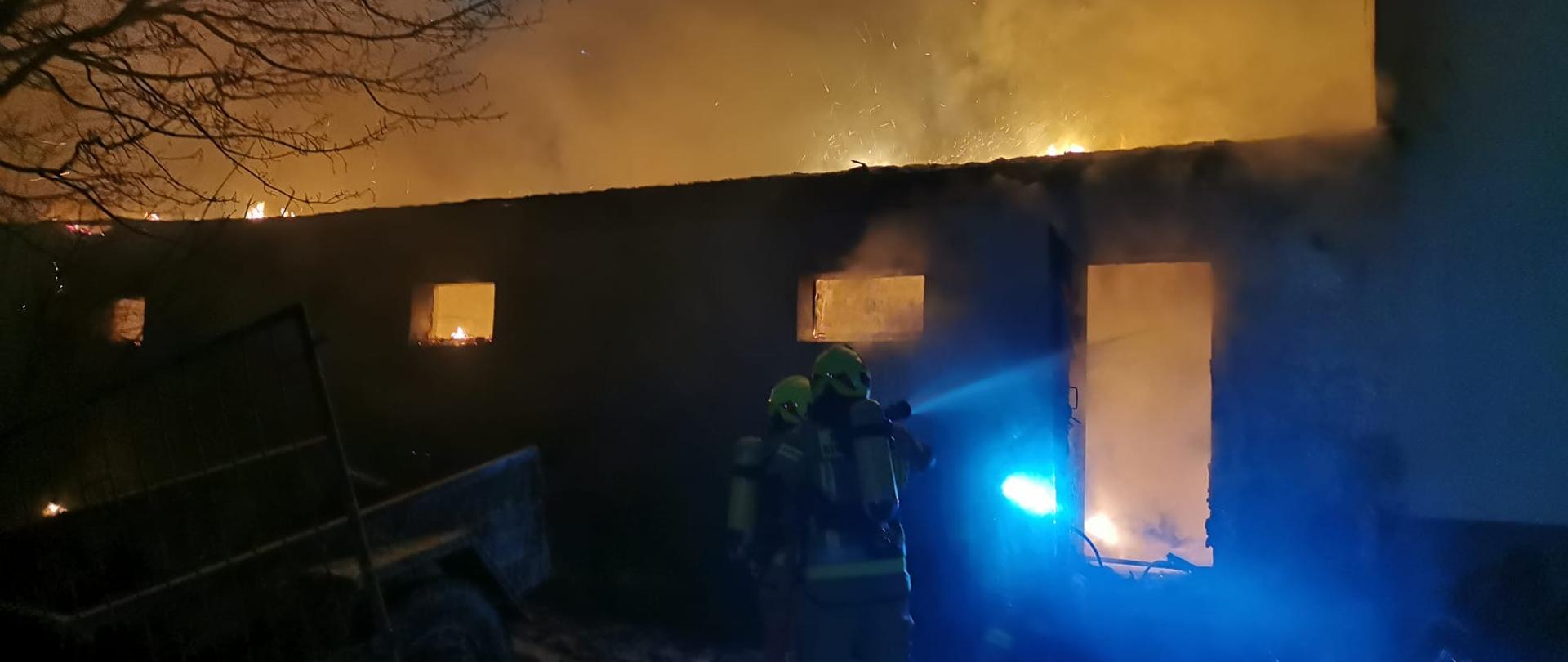 Parterowy budynek produkcyjno-magazynowy objęty pożarem, widoczne kłęby dymu.Przed budynkiem dwóch strażaków oświetlających wejście do budynku. Z prawej strony przylegający budynek mieszkalny.