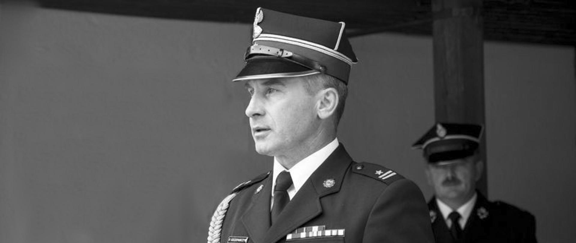 Zdjęcie czarno-białe. Na pierwszym planie ś.p. mł. bryg. w st. spocz. Paweł Szczepańczyk w mundurze wyjściowym PSP. W tle po prawej nieostra postać strażaka w mundurze wyjściowym OSP.