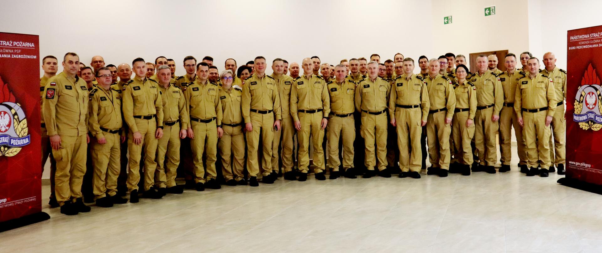 Duża grupa funkcjonariuszy PSP w mundurach stoi w sali, ustawiona do wspólnego zdjęcia. Po bokach znajdują się dwa roll-upy z logo PSP i napisem „Biuro Przeciwdziałania Zagrożeniom”.
