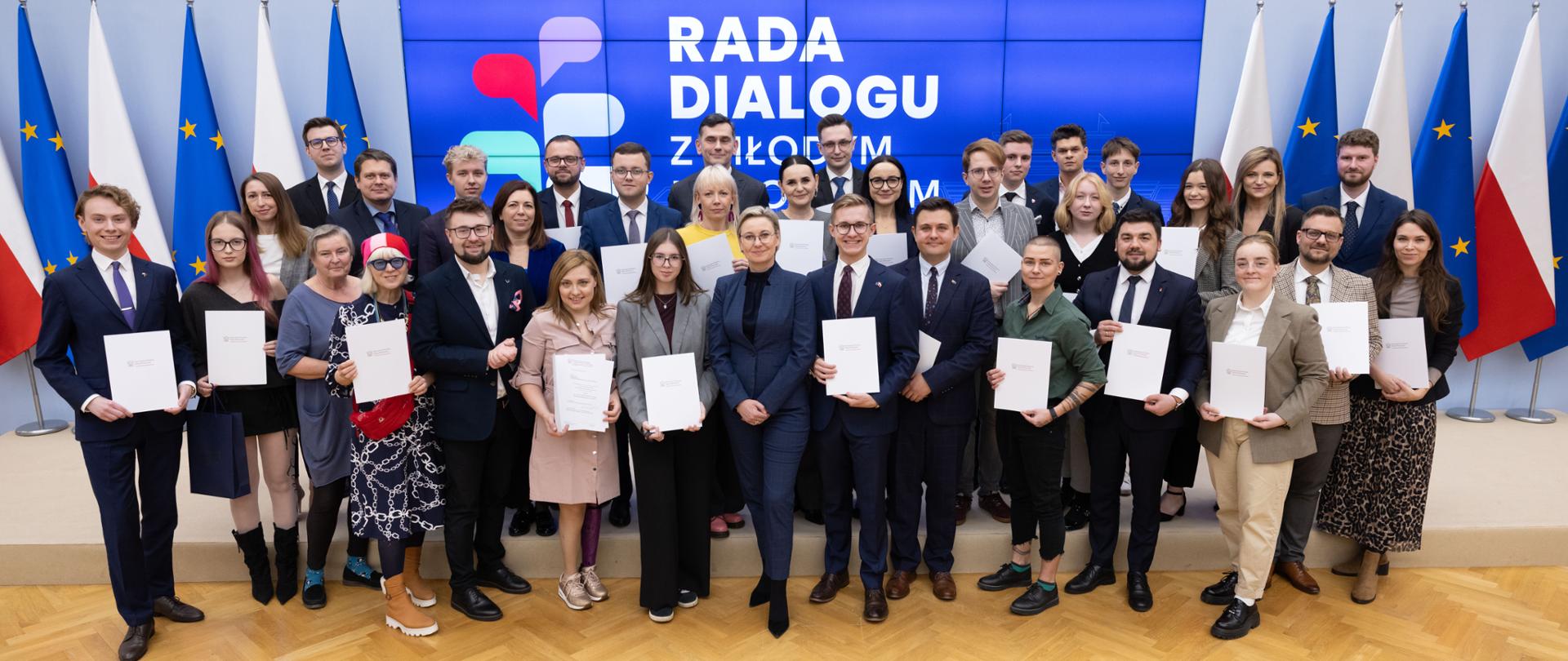 2025.10.30 Warszawa, Kancelaria Prezesa Rady Ministrów. I Posiedzenie Rady Dialogu z Młodym Pokoleniem IV kadencji. Zdjęcie grupowe na tle ekranu z logo i napisem Rada Dialogu z Młodym Pokoleniem. Na górze ekranu widoczne loga Kancelaria Prezesa rady Ministrów i Komitet do spraw Pożytku Publicznego. Po bokach ekranu widoczne są flagi Polski i Unii Europejskiej. fot. Stefan Maszewski