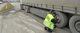 Umundurowany inspektor świętokrzyskiej Inspekcji Transportu Drogowego kontroluje stan techniczny naczepy ciężarowej. Funkcjonariusz sprawdza układ hamulcowy przy lewym kole na pierwszej osi naczepy.