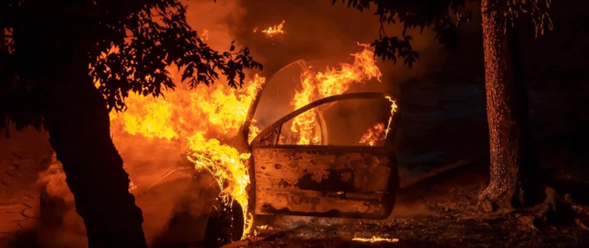 Na zdjęciu widoczny pojazd samochodowy, który jest całly w ogniu. Ogień wydobywa się spod pokrywy silnika oraz z otwartych drzwi kierowcy.