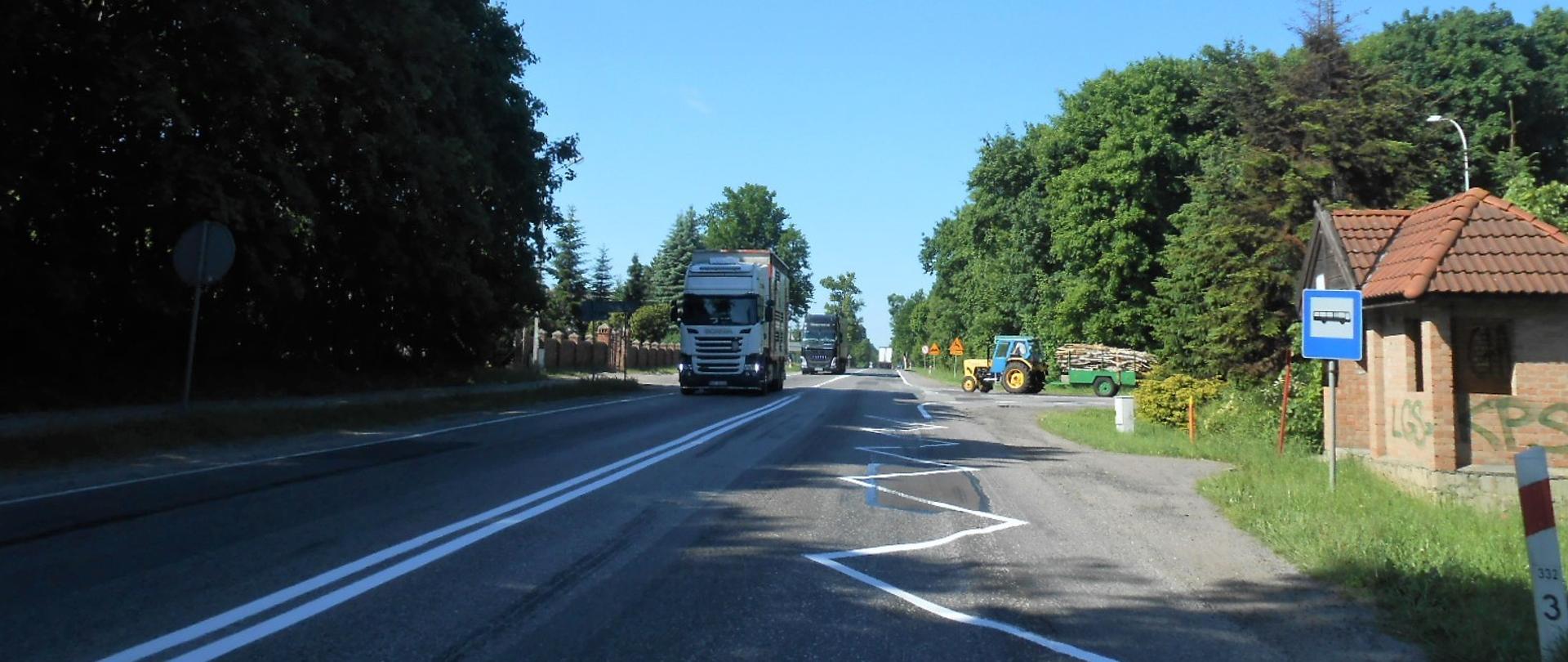 Zdjęcie przedstawia drogę krajową nr 22 w Swarożynie z jadącymi pojazdami. Na poboczu po prawej stronie widać przystanek komunikacji autobusowej pozbawiony zatoki autobusowej, a na dalszym planie ciągnik włączający się do ruchu z drogi podporządkowanej.