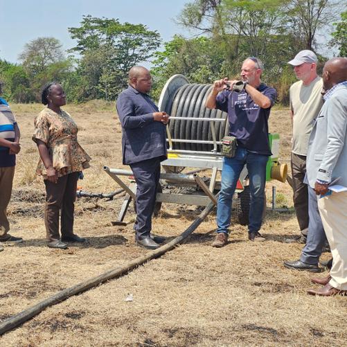 Mężczyzna coś opowiada i pokazuje rękoma, grupa osób mu się przygląda, stoją na wypalonej od słońca trawie, widać długi wąż ogrodowy