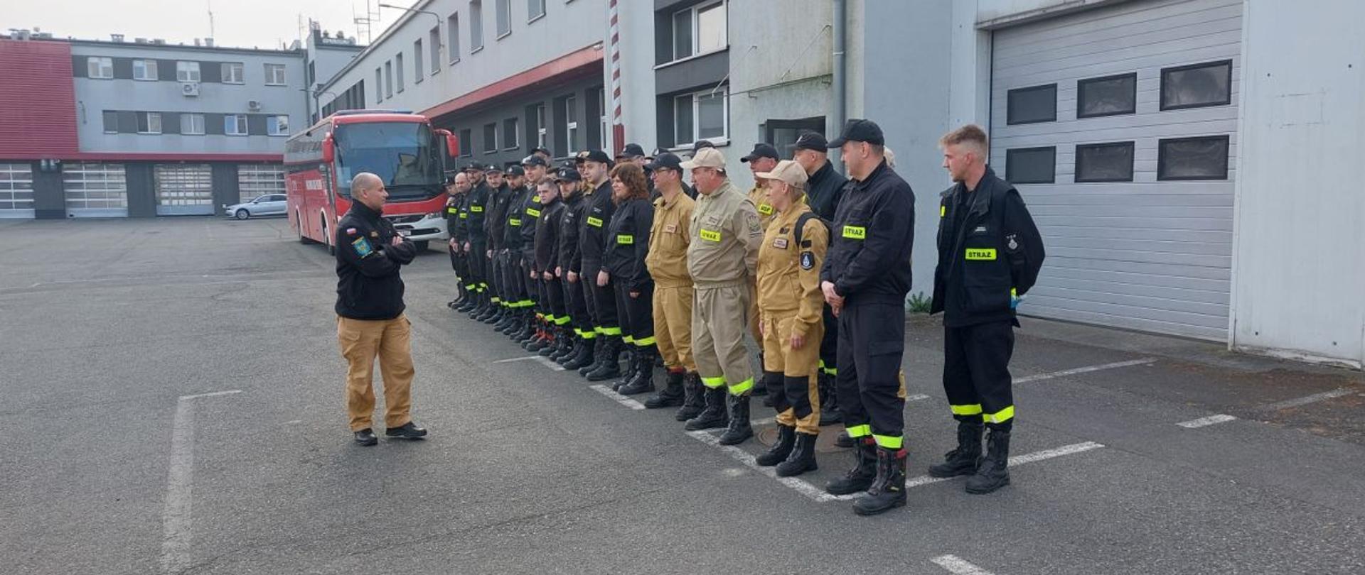 Grupa strażaków OSP w umundurowaniu koszarowym z napisem STRAŻ stoi w szeregu przed budynkiem Ośrodka Szkolenia KW PSP w Opolu. Przed nimi stoi prowadzący szkolenie, omawiający organizację dnia. W tle widoczny jest czerwony autobus oraz garaże jednostki. Scena przedstawia zbiórkę uczestników szkolenia kierujących działaniem ratowniczym.