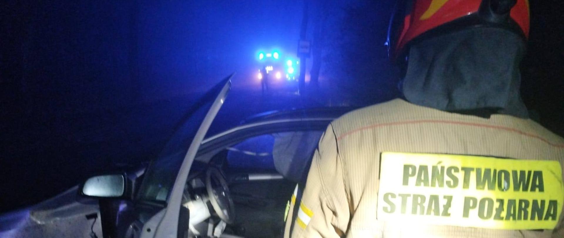 Zdjęcie zrobione nocą. Strażak stoi tyłem w mundurze bojowym czerwonym hełmie. na tle szarego samochodu osobowego. Mgła, w oddali niebieskie światła pojazdów ratowniczych.