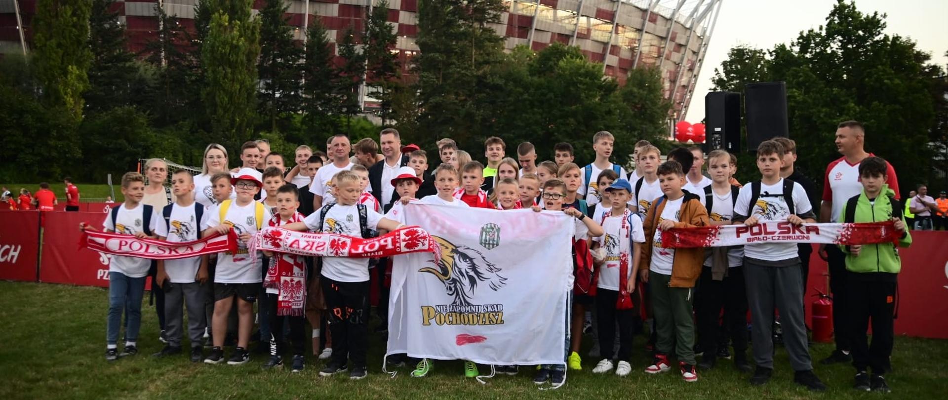 Na zielonej trawie stoi duża grupa dzieci trzymających biało-czerwone szaliki, za nimi drzewa i Stadion Narodowy.
