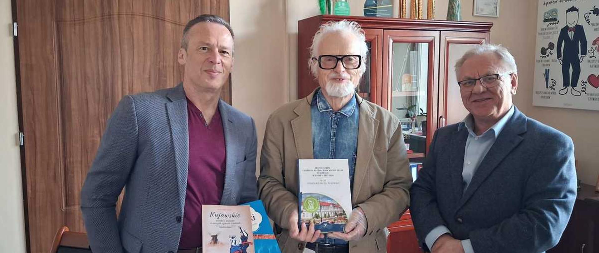 "Opis ogólny
Zdjęcie przedstawia trzech uśmiechniętych, elegancko ubranych mężczyzn stojących obok siebie w jasnym gabinecie. Mężczyźni prezentują do aparatu trzymane w dłoniach książki. Atmosfera spotkania jest oficjalna, a zarazem bardzo serdeczna.
Postacie (od lewej do prawej)
1. Mężczyzna po lewej stronie (Arkadiusz Ciechalski):
Jest to szczupły, wysoki mężczyzna w średnim wieku z krótkimi, ciemnymi włosami. Ma na sobie grafitową, lekko wzorzystą marynarkę, bordowy tiszert (lub cienki sweter) pod spodem oraz ciemne spodnie. W prawej ręce, na wysokości pasa, trzyma dwie książki o miękkich, kolorowych okładkach – jedna z nich ma widoczny duży napis „Kujawskie...”.
2. Mężczyzna w środku (Bogdan Lisowski):
To starszy, postawny mężczyzna z siwą, krótką brodą i gęstymi, zaczesanymi do góry siwymi włosami. Nosi charakterystyczne, wyraziste okulary w grubych, czarnych oprawkach. Ubrany jest w jasnobrązową (beżową) marynarkę, błękitną koszulę oraz jasne spodnie. Oburącz trzyma przed sobą pionowo białą książkę z kolorową grafiką przedstawiającą budynki szkoły oraz zielonym logo z liczbą na okładce.
3. Mężczyzna po prawej stronie (Dyrektor Andrzej Zdrojewski):
To nieco niższy starszy mężczyzna w okularach, o siwiejących włosach i przyjaznym wyrazie twarzy. Ma na sobie ciemnoniebieską marynarkę, jasnoniebieską koszulę z rozpiętym kołnierzykiem oraz ciemne spodnie. Stoi z dłońmi splecionymi na wysokości brzucha, uśmiechając się ciepło w stronę obiektywu.
Tło i otoczenie
Mężczyźni stoją w dobrze oświetlonym biurze (gabinecie dyrektora).
• Po lewej stronie w tle widoczne są zamknięte, brązowe, drewniane drzwi.
• Za plecami mężczyzn stoi wysoka, elegancka witryna z ciemnego drewna z przeszklonymi drzwiczkami, w której widać książki i dokumenty.
• Na szczycie szafy stoją pamiątki i nagrody: po prawej stronie duży, złoty puchar sportowy, pośrodku mniejsze statuetki, a po lewej stronie maskotka przedstawiająca uśmiechniętą, łaciatą krowę w zielonym stroju.
• Po prawej stronie, na jasnej ścianie, wisi pionowy plakat lub tablica pamiątkowa z rysunkową grafiką, nagłówkiem zaczynającym się od słów „Oto Andrzej dyrektor os. bud. ZSCKR w...” oraz mniejszymi ilustracjami i napisami."