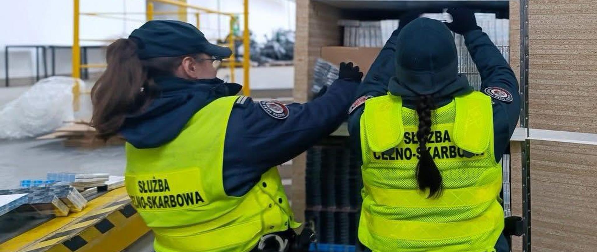 Dwie osoby w kamizelkach odblaskowych z napisem „Służba Celno‑Skarbowa” układają kartony i opakowania na półkach w magazynie. W tle widoczna jest hala magazynowa z regałami i sprzętem.