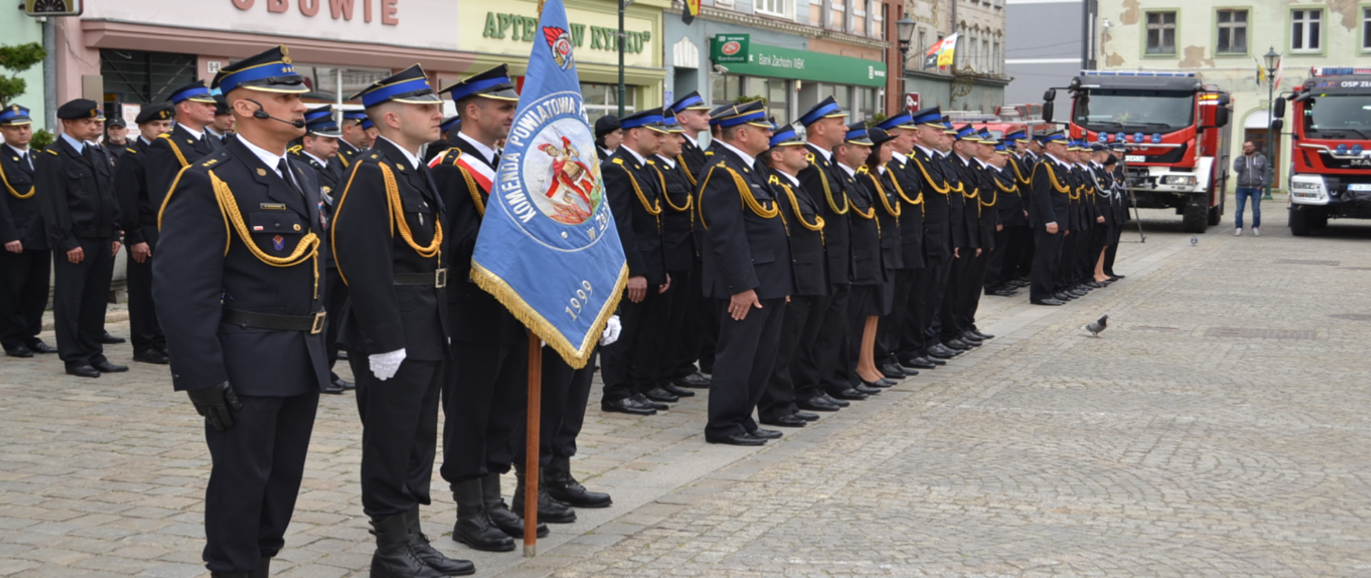 Zdjęcie przedstawia strażaków podczas uroczystości dnia strażaka na Żarskim rynku. Strażacy stoją w mundurach galowych.