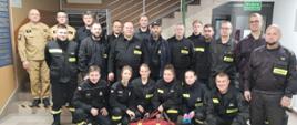 Na zdjęciu w holu Komendy Powiatowej Państwowej Straży Pożarnej w Gorlicach strażacy z Ochotniczych Straży Pożarnych i członkowie Komisji egzaminacyjnej strażacy z Państwowej Straży Pożarnej po egzaminie kończącym kurs z zakresu kwalifikowanej pierwszej pomocy i recertyfikacji uprawnień dla druhów OSP z powiatu gorlickiego. Strażacy ubrani w ubrania koszarowe koloru czarnego i piaskowego bez czapek.