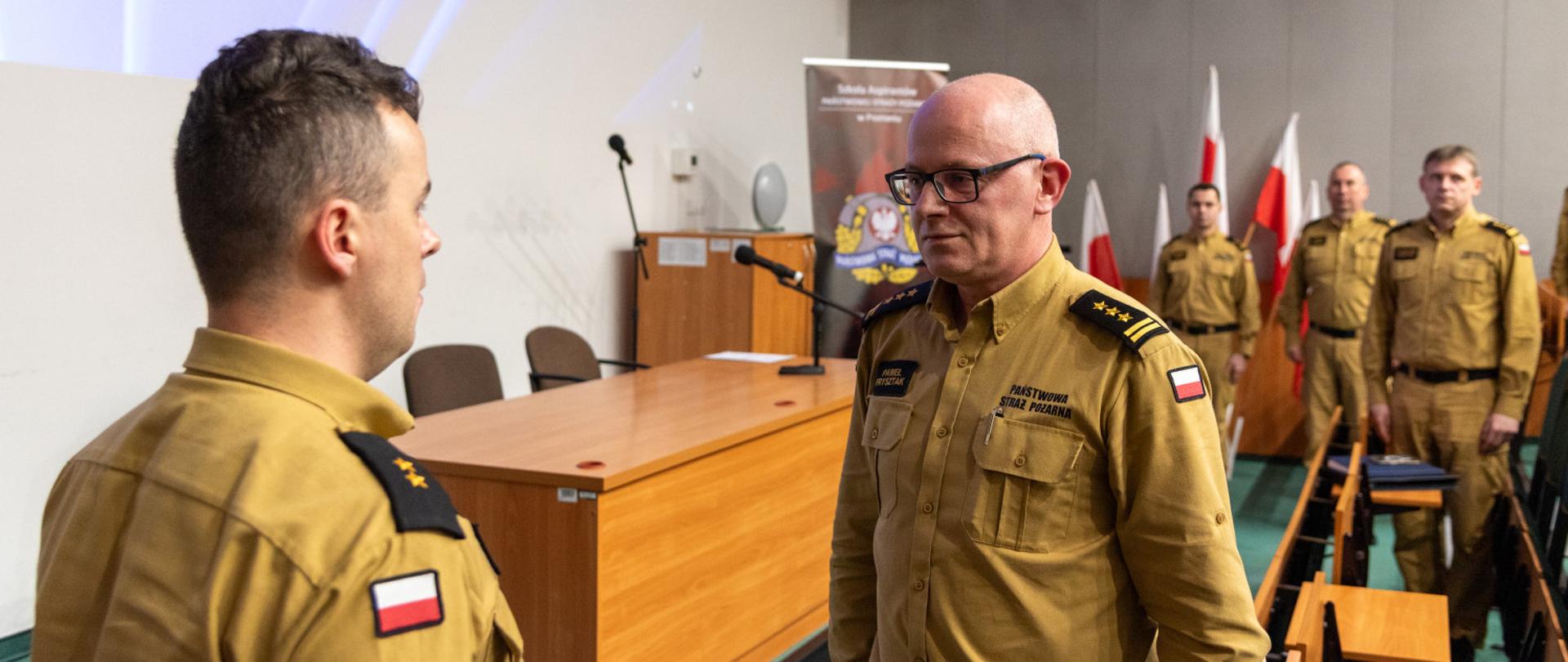Na zdjęciu znajduje się grupa mężczyzn w mundurach strażackich w sali wykładowej. Dwóch mężczyzn na pierwszym planie prowadzi rozmowę – jeden z nich, starszy mężczyzna w okularach, trzyma w ręku teczkę, a drugi, młodszy, stoi naprzeciwko niego. W tle widać kilku innych funkcjonariuszy w takich samych mundurach oraz polskie flagi ustawione przy ścianie. Na ścianie znajduje się ekran z niebieską grafiką, a przed nim stoi drewniane biurko z mikrofonem. Zdjęcie sugeruje oficjalne spotkanie w jednostce Państwowej Straży Pożarnej.