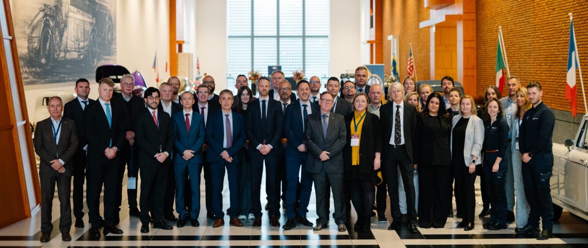 Wspólne zdjęcie uczestników międzynarodowego spotkania w reprezentacyjnej sali z wysokim, łukowym sufitem. Grupa około 40 osób stoi na środku jasnego holu, a w tle widoczne są flagi kilku państw oraz ekspozycja muzealna.