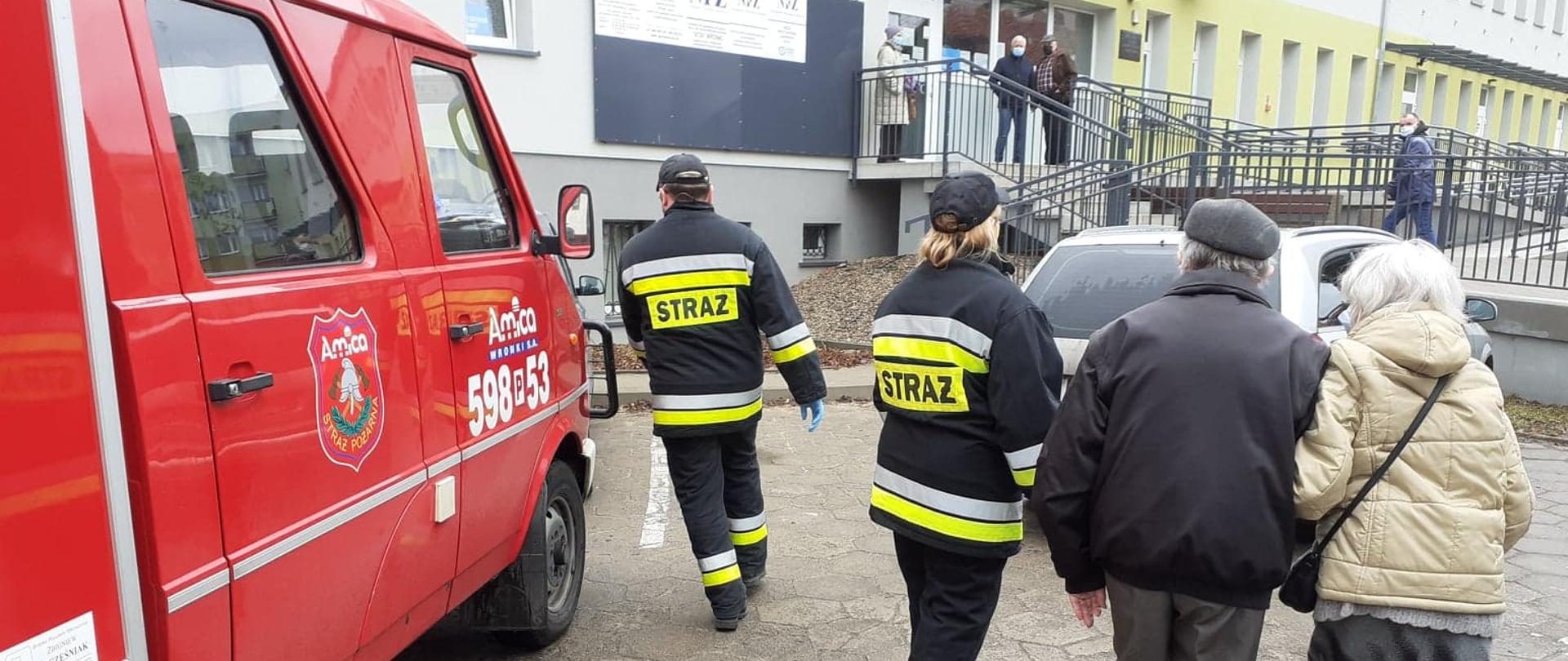 Widać strażaków i dwie osoby, które udają się do punktu szczepień