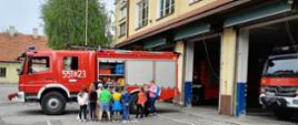 Widok z boku. Dzieci z opiekunką i strażak stoją obok samochodu strażackiego przed garażami JRG. Strażak prezentuje wyposażenie pojazdu. 