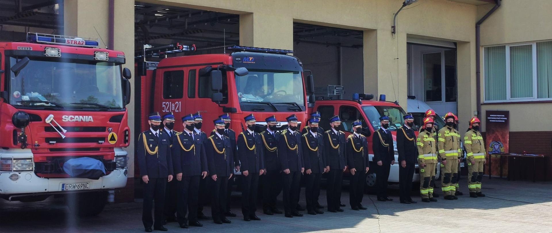 Zdjęcie przedstawia plac przed budynkiem komendy, wyłożony kostką i stojących przed nim, w dwuszeregu strażaków, w galowym oraz bojowym umundurowaniu, a za nimi stoją samochody strażackie w otwartych drzwiach garażowych strażnicy.