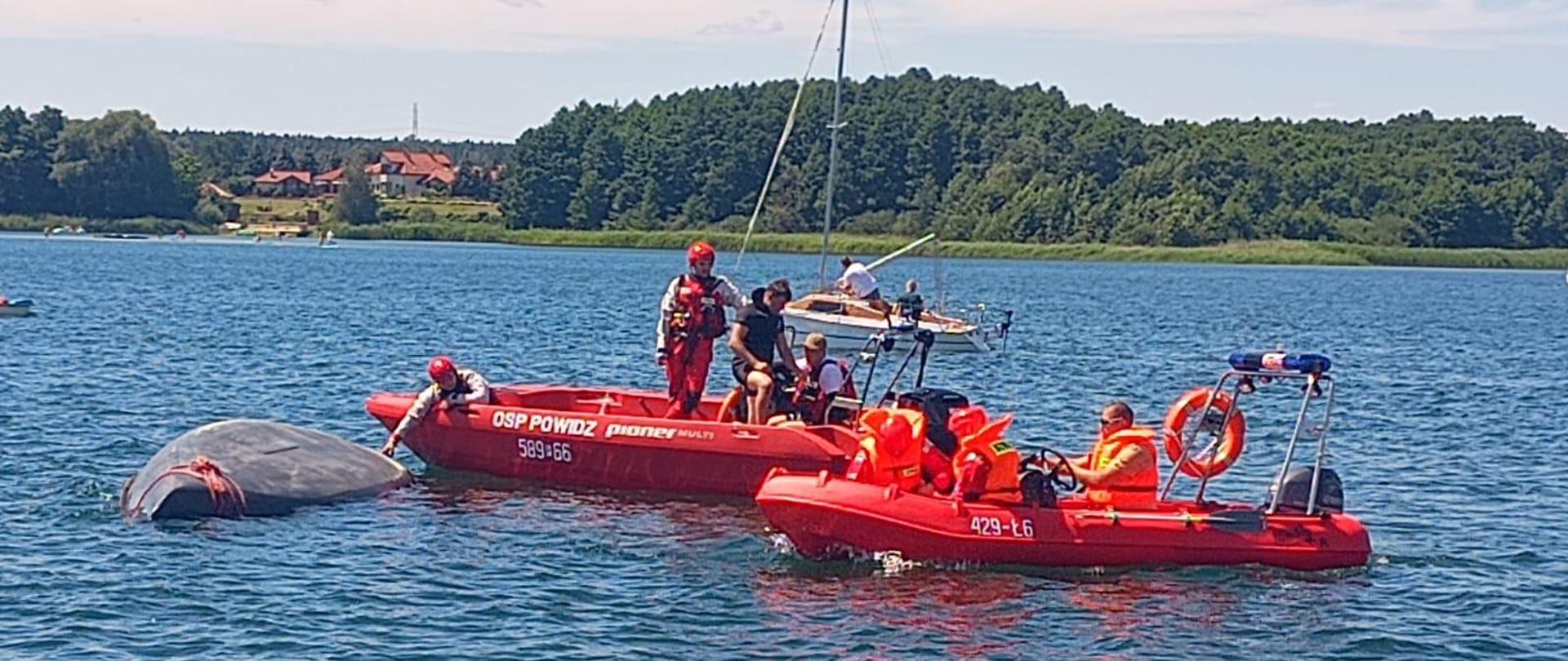 Na zdjęciu widać jak strażacy próbują podnieść żaglówkę by bezpiecznie z holować ją do brzegu. Maszt żaglówki dotyka dna.