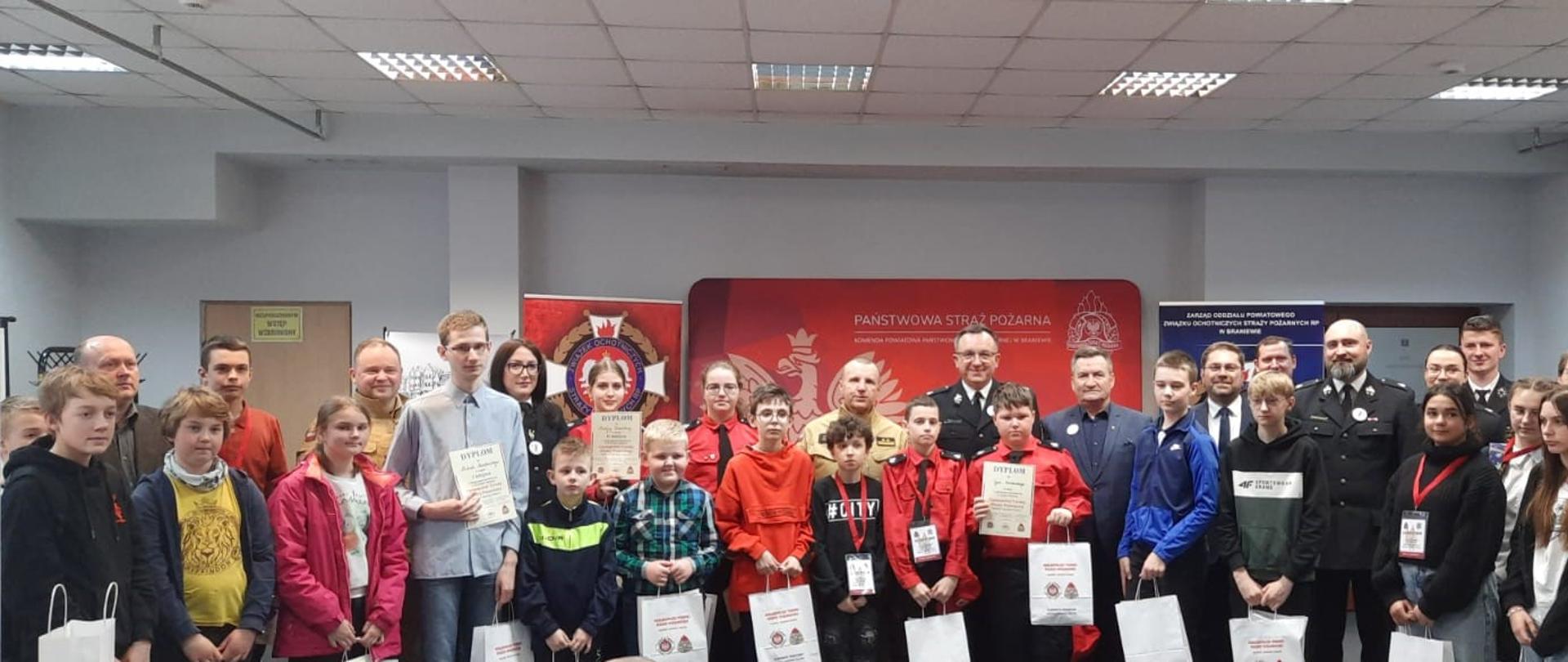 Na zajęciu duża grupa dzieci, uczestników konkursu. W rekach trzymają dyplomy i upominki. Za dziećmi stoją organizatorzy konkursu.