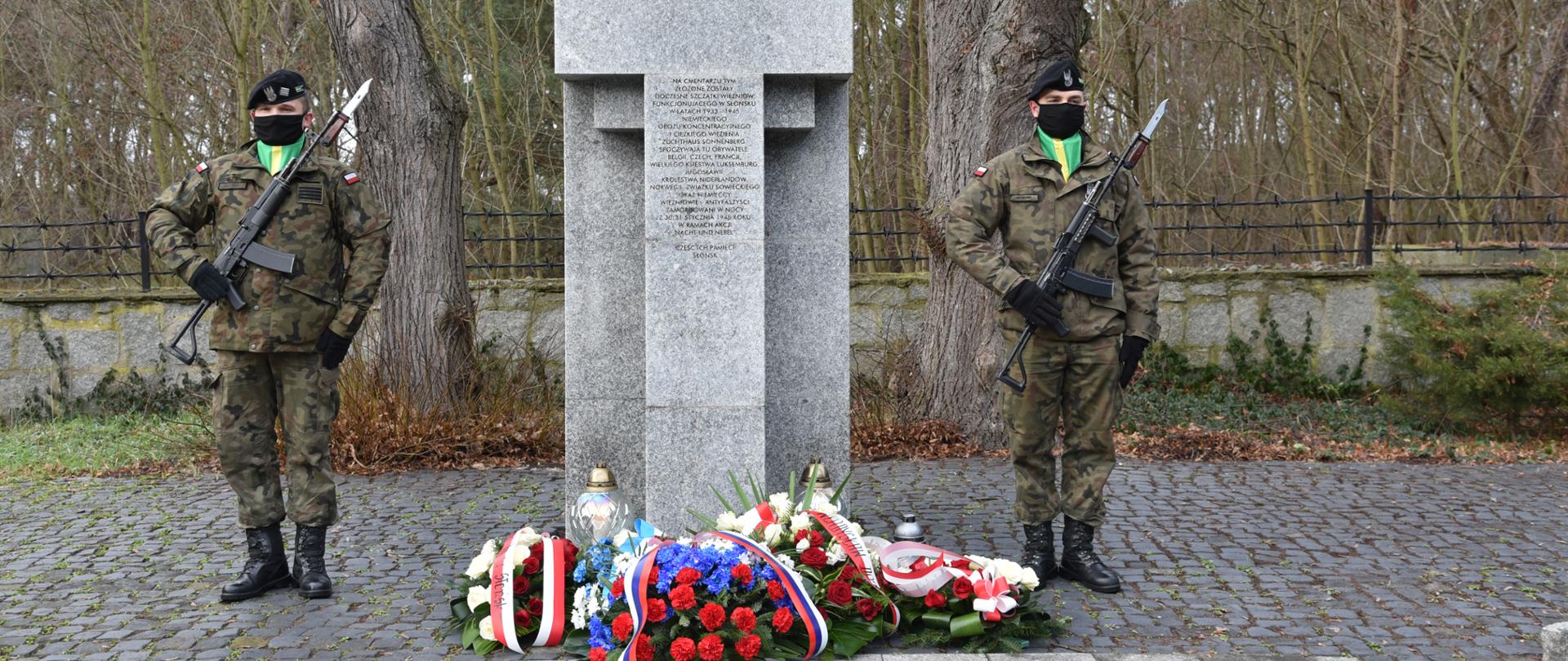 Na środku kamienny pomnik w kształcie krzyża. Po jego prawej i lewej stronie stoi żołnierz. Pod pomnikiem leżą złożone wieńce i stoją zapalone znicze